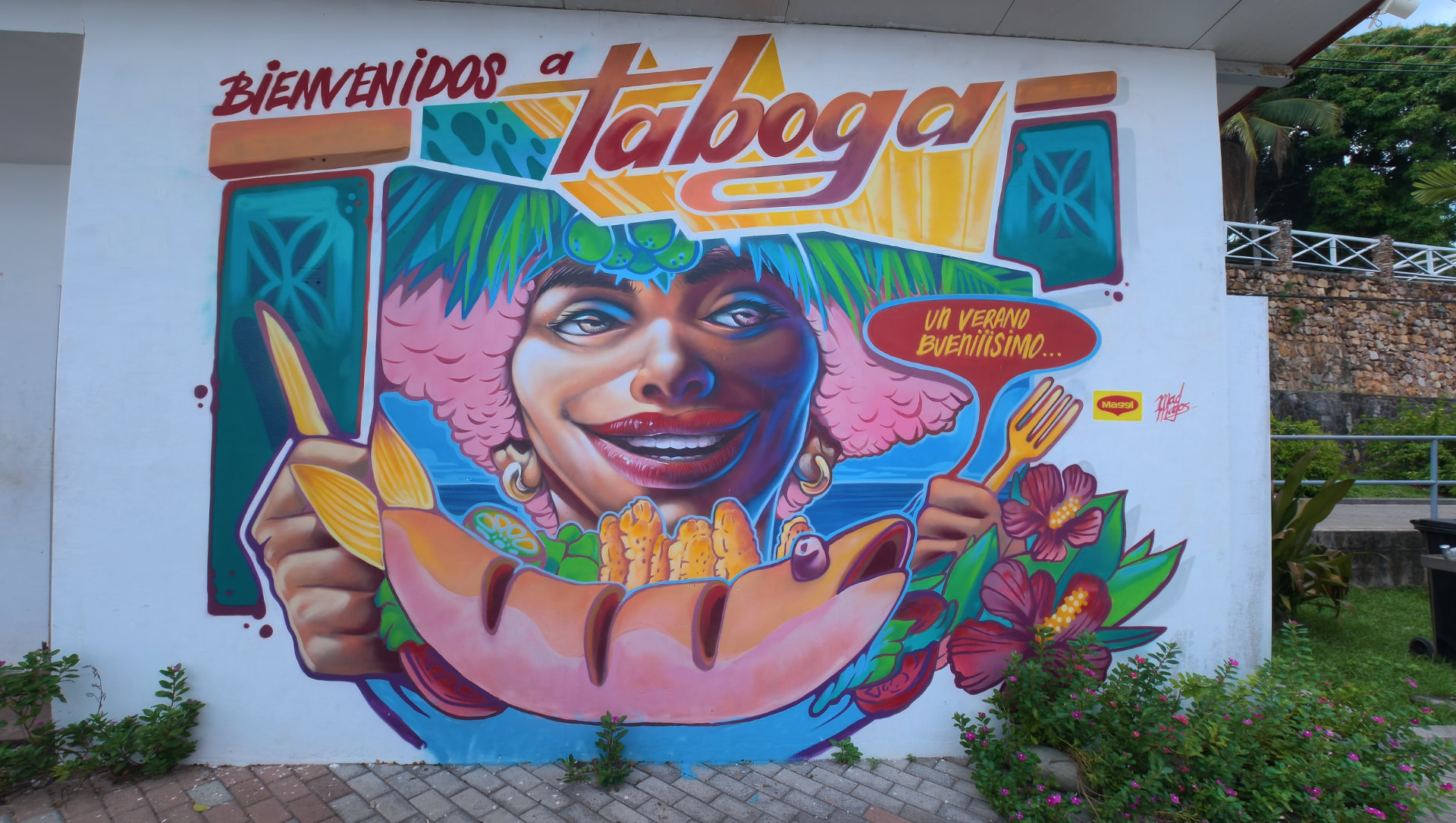 Mural 'Bienvenidos a Taboga' en una pared blanca del pueblo: una mujer sonriente con tocado de hojas tropicales frente a un plato de pescado
