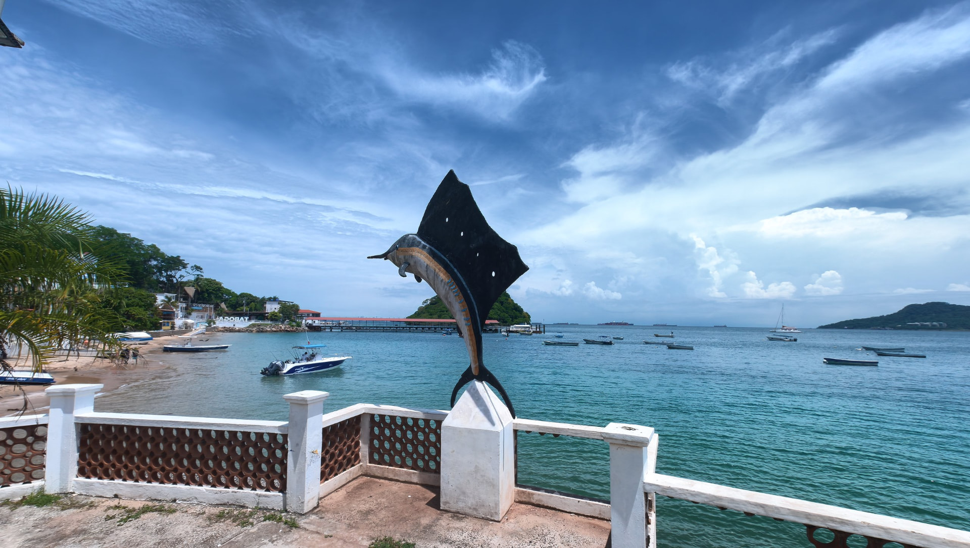 Escultura de pez vela sobre un pilar blanco en el mirador, con la bahía turquesa, la playa y el letrero de Taboga al fondo